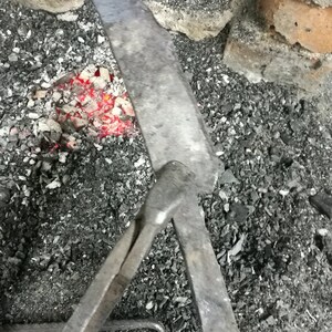 May include: A blacksmith's tool with a long handle and a curved hook at the end. The tool is resting on a bed of hot coals.