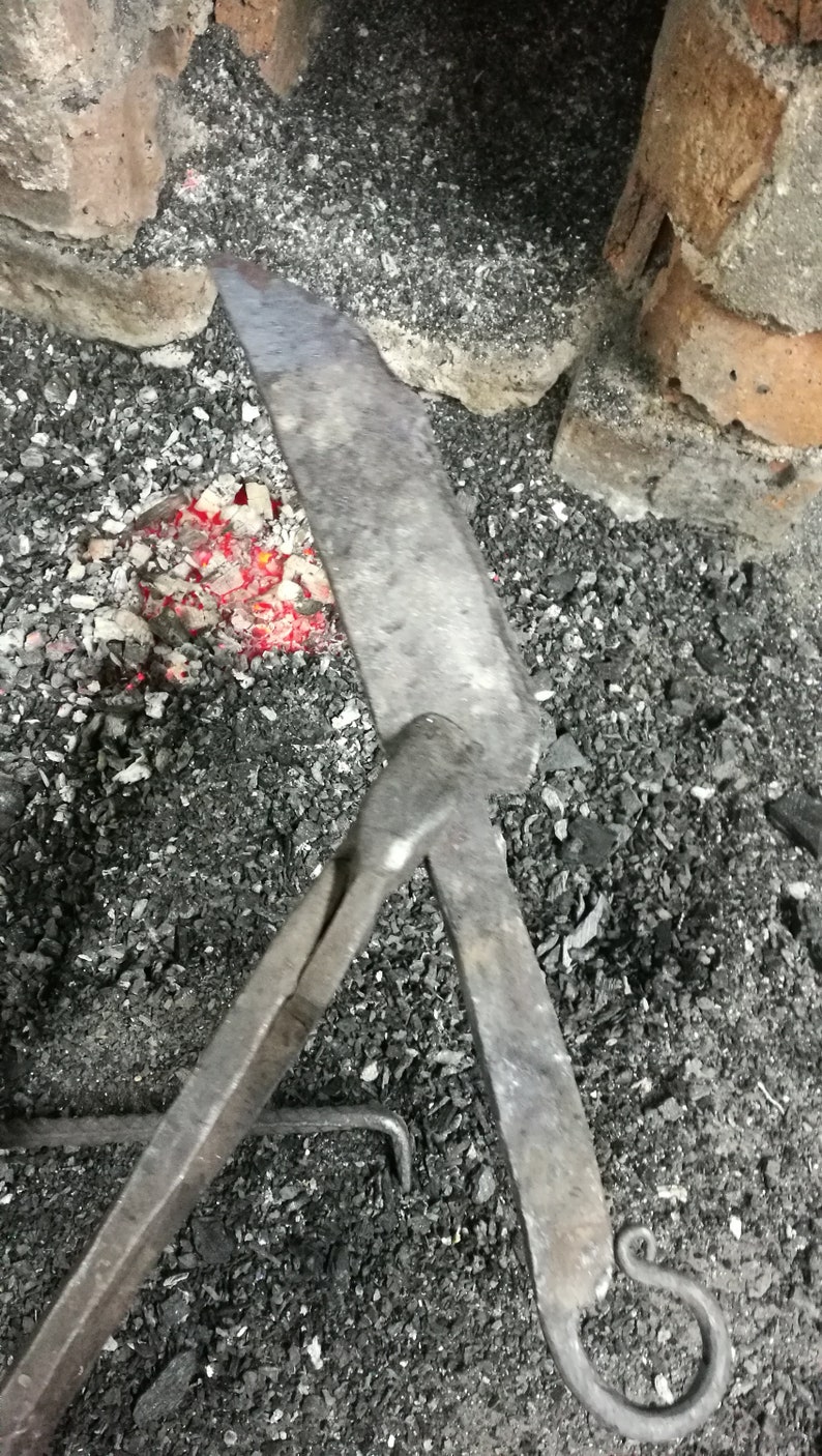 May include: A blacksmith's tool with a long handle and a curved hook at the end. The tool is resting on a bed of hot coals.