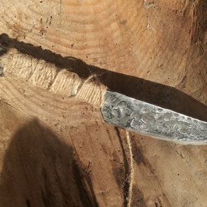 May include: A hand-forged knife with a rustic, hammered finish. The blade is wrapped with twine and has a metal ring at the end of the handle.