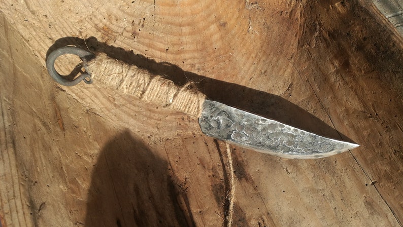 May include: A hand-forged knife with a rustic, hammered finish. The blade is wrapped with twine and has a metal ring at the end of the handle.