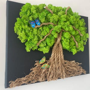 May include: A decorative wall art piece featuring a tree design. The tree's foliage is made of vibrant green moss, with a trunk crafted from rope. Two blue butterflies and one multi-colored butterfly are attached to the tree.