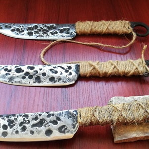 May include: Three handmade knives with hammered steel blades and wrapped rope handles. The knives have a rustic, primitive look.