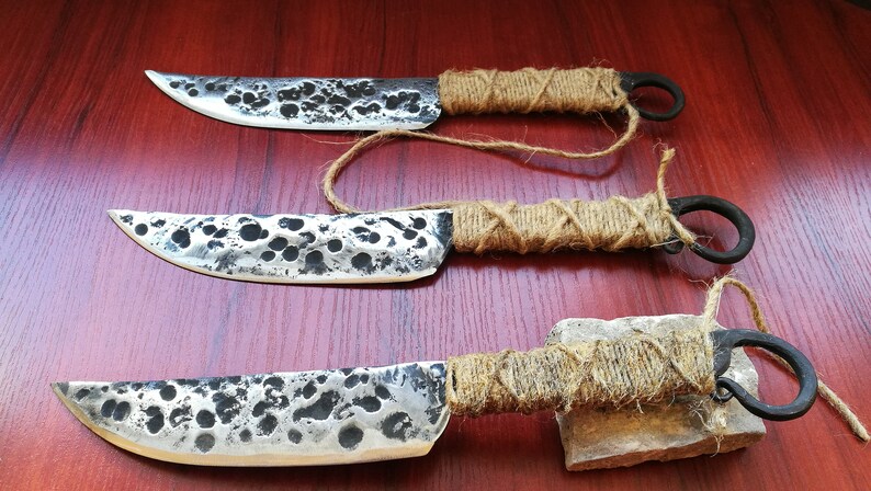 May include: Three handmade knives with hammered steel blades and wrapped rope handles. The knives have a rustic, primitive look.