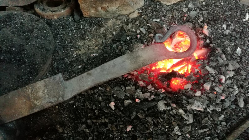 May include: A metal rod is heated in a forge with glowing red hot coals. The rod has a curved end that is being shaped into a hook.