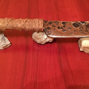 May include: A handmade knife with a rustic, hammered finish. The blade is made of metal and has a brown patina. The handle is wrapped in twine and has a metal loop at the end.
