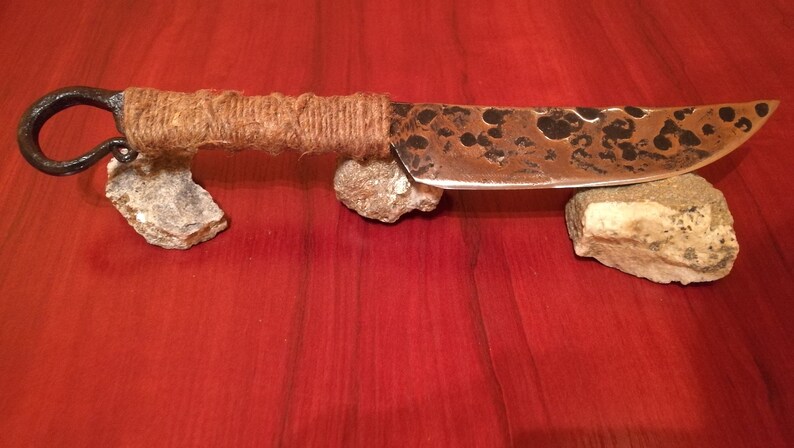 May include: A handmade knife with a rustic, hammered finish. The blade is made of metal and has a brown patina. The handle is wrapped in twine and has a metal loop at the end.