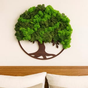 Peut inclure: Une décoration murale ronde en forme d'arbre. Le feuillage de l'arbre est composé de mousse verte vive, contrastant avec un tronc et des branches en bois brun foncé. L'œuvre est fixée sur un mur blanc au-dessus d'une tête de lit et d'oreillers en bois.