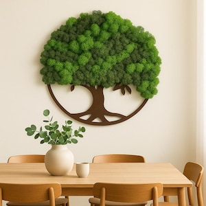 Può includere: Un'opera d'arte da parete rotonda in legno marrone con un albero della vita e una chioma verde strutturata simile al muschio. L'opera è montata su una parete bianca sopra un tavolo da pranzo in legno. Un vaso bianco con foglie verdi e una tazza bianca sono sul tavolo.