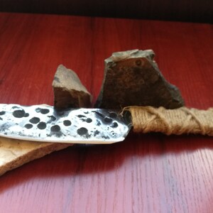 May include: A handmade knife with a hammered steel blade and a brown rope handle. The knife is resting on a piece of stone.