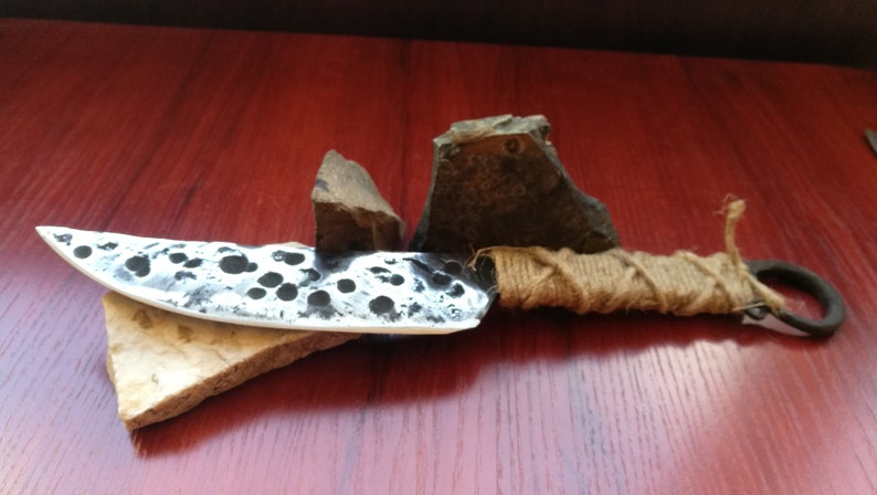 May include: A handmade knife with a hammered steel blade and a brown rope handle. The knife is resting on a piece of stone.