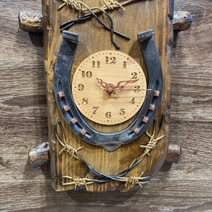 May include: A rustic wooden clock with a horseshoe design and a clock face with Roman numerals. The clock is surrounded by barbed wire.