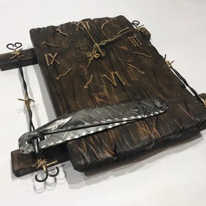 Könnte beinhalten: Eine rustikale Holzuhr mit römischen Ziffern und goldfarbenen Zeigern. Die Uhr ist an einer Metallplatte mit Stacheldrahtdesign befestigt. Die Uhr ist auf einem Holzbrett mit einem Metallscharnier montiert.