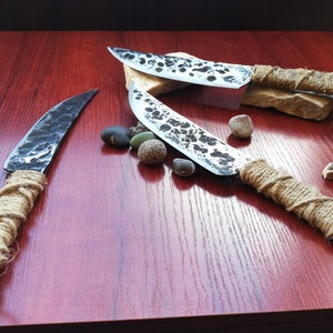 May include: Three handmade knives with rustic, hammered blades and wrapped rope handles. The knives are displayed on a wooden surface with small stones.