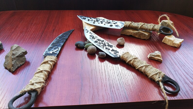 May include: Three handmade knives with rustic, hammered blades and wrapped rope handles. The knives are displayed on a wooden surface with small stones.