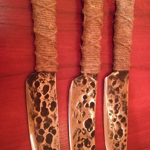 May include: Three handmade knives with brown rope handles and hammered metal blades. The blades have a rustic, hammered finish.