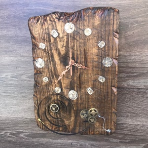 May include: A rustic wooden clock with a dark brown stain. The clock face is made of metal gears and the hands are copper coloured. The clock is hanging on a wooden wall.