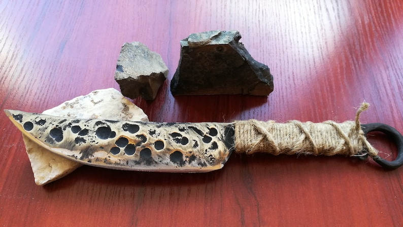 May include: A handmade knife with a hammered, textured blade and a brown rope handle. The knife is resting on a white rock and there are two gray rocks in the background.