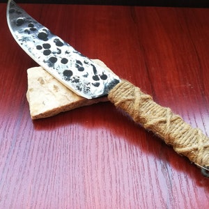 May include: A handmade knife with a black and white speckled blade and a brown rope handle. The handle is wrapped in a criss-cross pattern and has a metal ring at the end.