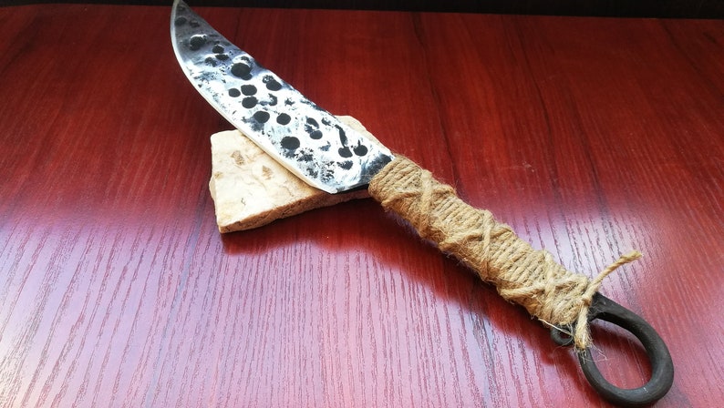 May include: A handmade knife with a black and white speckled blade and a brown rope handle. The handle is wrapped in a criss-cross pattern and has a metal ring at the end.