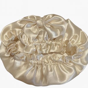 May include: A cream-colored satin bonnet with a large bow on top. The bonnet has a ruffled edge and a gathered elastic band. The fabric has a shiny, smooth texture. This hair accessory is designed to protect hair while sleeping.