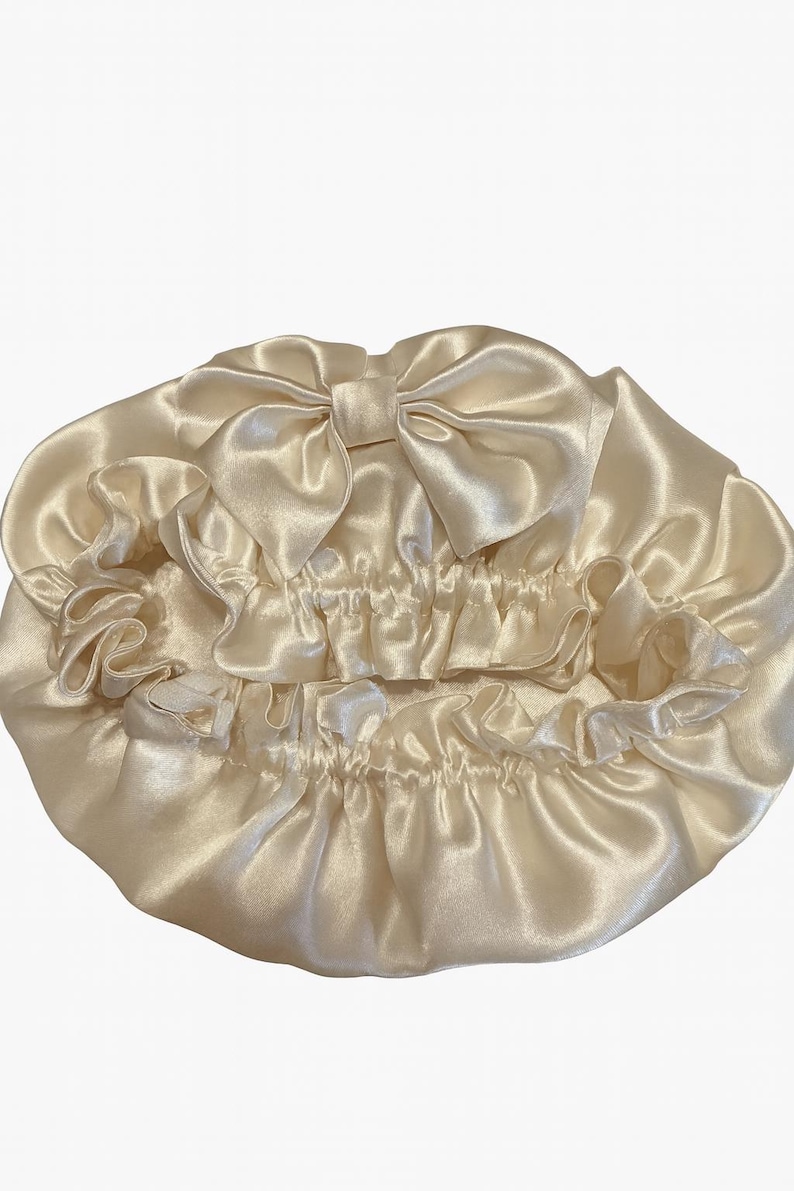 May include: A cream-colored satin bonnet with a large bow on top. The bonnet has a ruffled edge and a gathered elastic band. The fabric has a shiny, smooth texture. This hair accessory is designed to protect hair while sleeping.