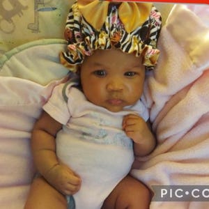 May include: A baby wearing a bonnet with a zebra print design, with a pink and gold color scheme. The baby is wearing a light pink onesie and is lying on a pink blanket.