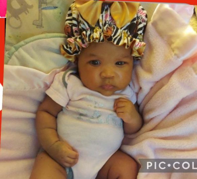 May include: A baby wearing a bonnet with a zebra print design, with a pink and gold color scheme. The baby is wearing a light pink onesie and is lying on a pink blanket.