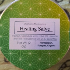 May include: A white label with green and white geometric pattern on a silver tin container. The label reads "Moshier's Mushrooms Healing Salve Plantain Calendula, Violet. Olive Oil Lavender and Tea Tree Essential Oil Vit E. Bees Wax Net Wt. (2 Oz) Homegrown, Foraged, Organic"