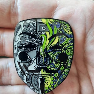 May include: A black and white enamel pin with a stylized face design. One half of the face is a detailed, intricate pattern of black and white lines. The other half of the face is a colorful pattern of green, yellow, and blue lines.