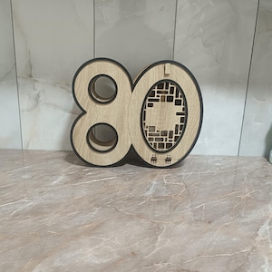 Pode incluir: Decoração de madeira com o número 80, com acabamento castanho claro e contornos pretos. O número tem dois recortes circulares no zero e um design decorativo recortado. O item é exibido em uma superfície com padrão de mármore.