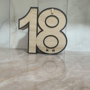 Könnte beinhalten: Holzzahl "18" in hellem Holzmaserungs-Finish mit schwarzem Rand. Die Zahl ist freistehend und hat ein dreidimensionales Aussehen. Die "8" hat zwei kreisförmige Aussparungen. Das Design ist schlicht und modern.
