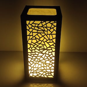 May include: A black lamp with a geometric pattern cut out of the sides. The lamp is lit and casting a warm yellow glow.