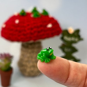 May include: A small, bright green frog figurine with black eyes and yellow accents. The frog is perched on a fingertip, with a blurred background featuring a red mushroom and a small plant.