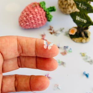 May include: A tiny, translucent butterfly with white and light blue wings rests on a finger. Scattered around are other miniature butterflies in various colors. A pink crocheted strawberry and a decorative piece with the words "Live Here" are in the background.