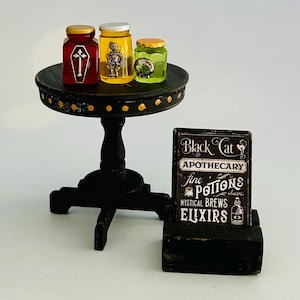 May include: Miniature Halloween apothecary scene featuring three glass jars with spooky designs on a small black table. A sign reads "Black Cat Apothecary" with text about potions and elixirs. The jars are filled with colorful liquids.