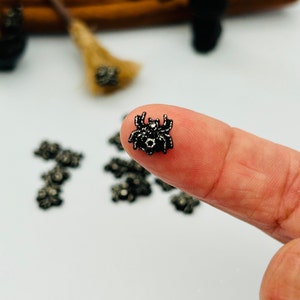 May include: A small, black spider charm with intricate detailing is held between fingers. The spider has a textured body and legs, with a small, round head. Other similar charms are scattered on a white surface. A miniature broom is in the background.