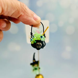 May include: A miniature black cauldron with a skull design, filled with green beads and blue accents, is suspended by a black handle. A small witch figure with a gold apple base hangs below. A yellow stirrer is in the cauldron.