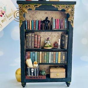 May include: Miniature black bookshelf with gold accents, filled with tiny books, a witch figurine, and other decorative items. A small pot with a plant and a crescent moon with a figurine are on top. The word "MAGIC" is visible.