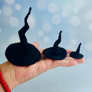May include: Three black witch hats of varying sizes, with a curved, pointed top and a wide brim. The hats are made of a soft, textured material. The hats are displayed on a hand.