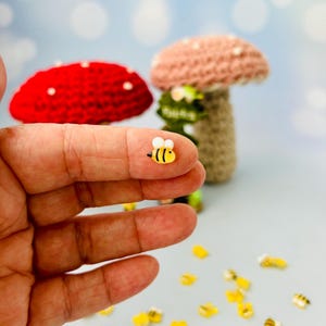 May include: A tiny, yellow and black bee-shaped embellishment is held in a person's hand. In the background, two crocheted mushrooms in red and pink are visible. Scattered around are more bee-shaped embellishments. The scene has a whimsical, handmade aesthetic.