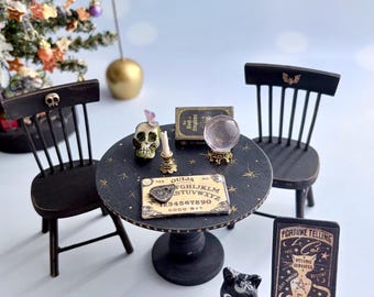 May include: A miniature black round table with gold star accents, two black chairs, a skull, a crystal ball, a Ouija board, a book, and a black cat figurine. A black sign reads Fortune Telling Cat, Psychic Services, 5, Always Open, Reveals The Future, No Refunds.