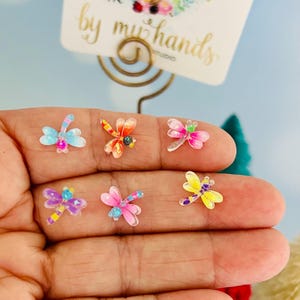 May include: Six small, colorful dragonfly-shaped charms featuring translucent wings and bodies in shades of blue, orange, pink, purple, and yellow. Each charm has a detailed, multi-colored pattern on the wings and a small, rounded head.