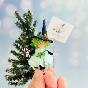 May include: A miniature witch doll with a black hat adorned with red berries and a green dress. The doll holds a candy cane and has green hair. A small Christmas tree is in the background. A card with text is also in the background.