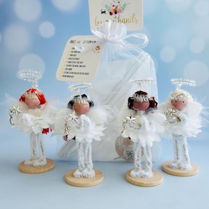 May include: Four handmade angel figurines with white feather wings, silver halos, and star accents. Each angel has a different hair color and stands on a wooden base. A clear bag with a tag that says "by hands" is in the background.