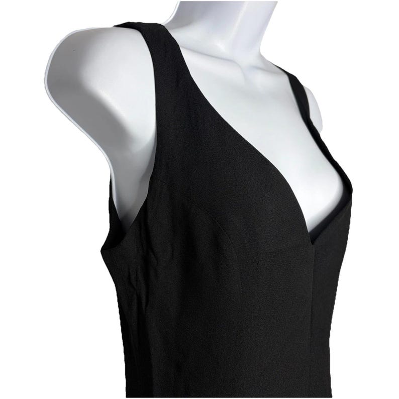 Op de afbeelding: Een zwarte mouwloze jurk met een diepe V-hals. De jurk is gemaakt van een textuurstof en heeft een eenvoudig, elegant ontwerp. De jurk wordt gepresenteerd op een witte mannequin.