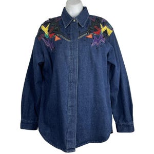 Może przedstawiać: Granatowa koszula dżinsowa z długimi rękawami. Koszula posiada kolorowe geometryczne wzory na ramionach, w tym żółte, czerwone i fioletowe kształty, ozdobione czarnymi koralikami. Koszula ma zapięcie na guziki i klasyczny kołnierz.