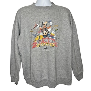 Puede incluir: Una sudadera gris jaspeado de manga larga y cuello redondo con un gráfico colorido de Mickey Mouse, Goofy, Pluto y el Pato Donald celebrando. El texto Happy New Year 2000 aparece junto al logotipo de Disneyland impreso bajo los personajes.