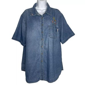 Könnte beinhalten: Hellblaues Jeanshemd mit kurzen Ärmeln. Das Hemd hat gestickte Blumen- und Vogelmotive auf der Brust und eine kleine Tasche. Das Hemd hat einen klassischen Kragen und Knopfverschlüsse vorne.