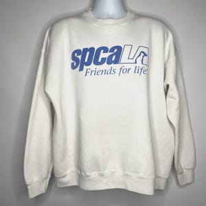 May include: A white crew neck sweatshirt with the blue text "spcaLA" and "Friends for life." The sweatshirt has long sleeves and a relaxed fit. The SPCA logo is also in blue.
