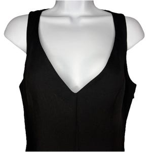 Op de afbeelding: Een zwarte mouwloze jurk met een diepe V-hals. De jurk wordt getoond op een witte mannequin torso, waarbij de snit en stijl worden benadrukt. De stof heeft een effen, matte uitstraling.